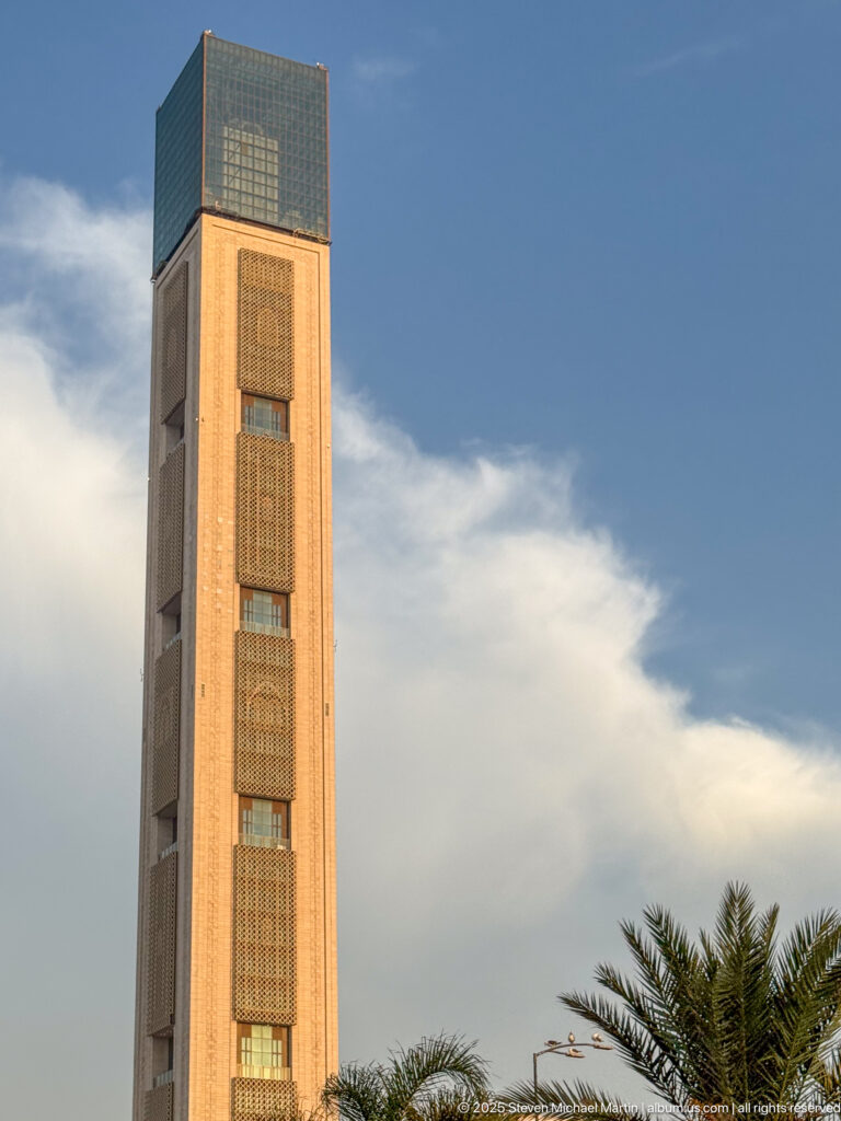 Close view of a tall rectangular tower with repeated patterned panels and a dark glazed cap against blue sky.