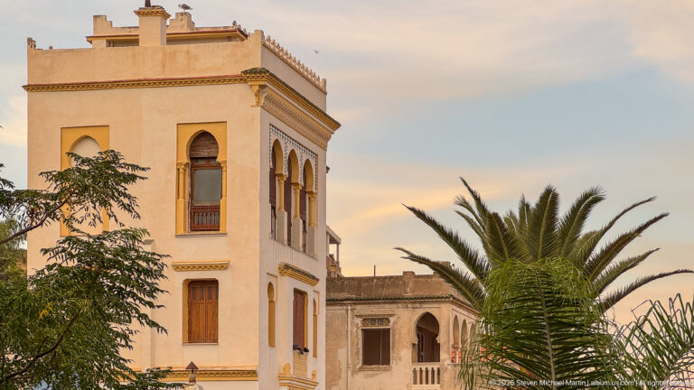 Cream-colored building with arched windows, decorative trim, corner tower, palms, and trees in Algiers.