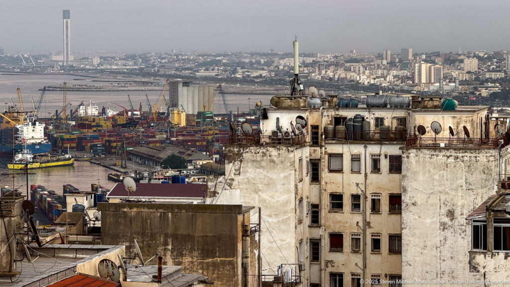Elevated view over weathered rooftops to Algiers harbor with containers, cranes, ships, and dense city beyond.