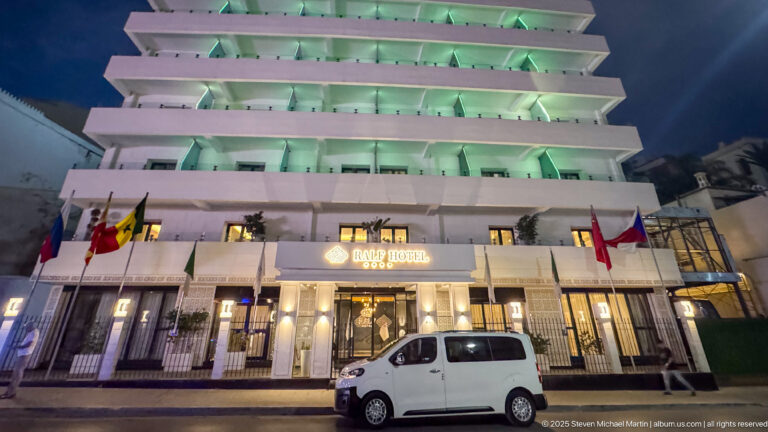 Night view of a multi-story hotel with stacked balconies, illuminated entrance, flags, and a white van at the curb.