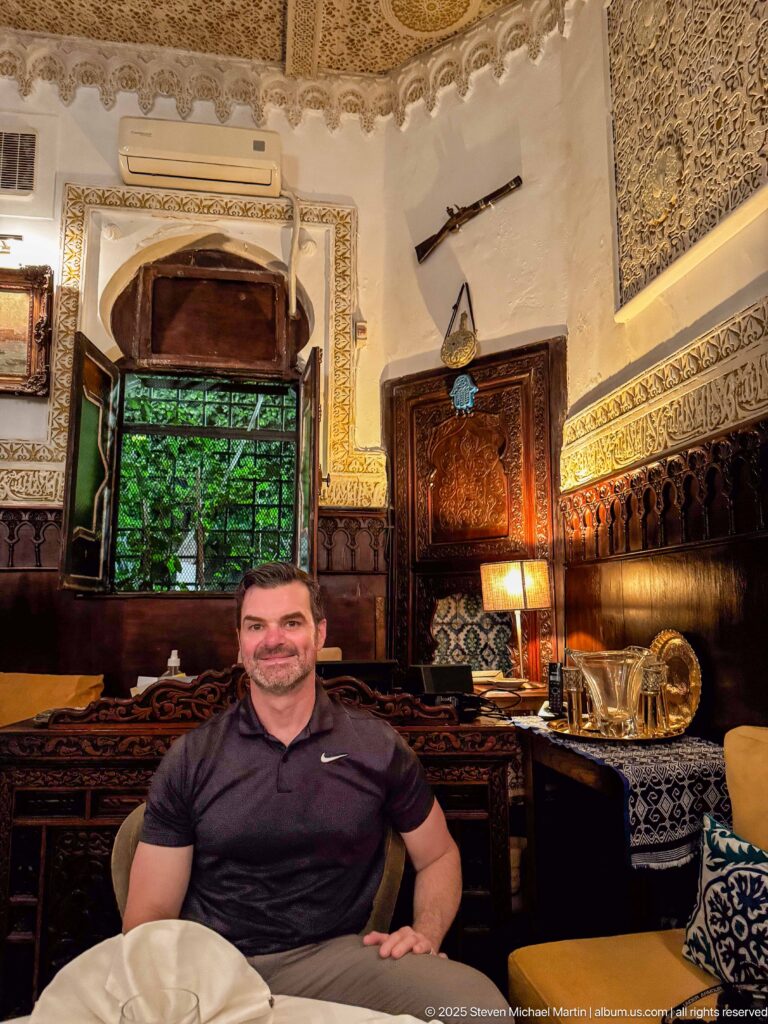Man seated in an ornate dining room with carved woodwork, patterned walls, shuttered window, and brass table service.