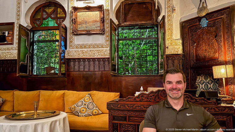 Man seated in an ornate Algiers dining room with carved woodwork, patterned walls, framed art, and mustard banquettes.