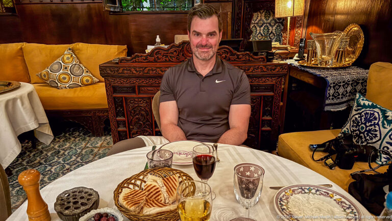 Man seated at a restaurant table with flatbread, glasses, patterned plates, mustard banquettes, and carved wood furnishings.