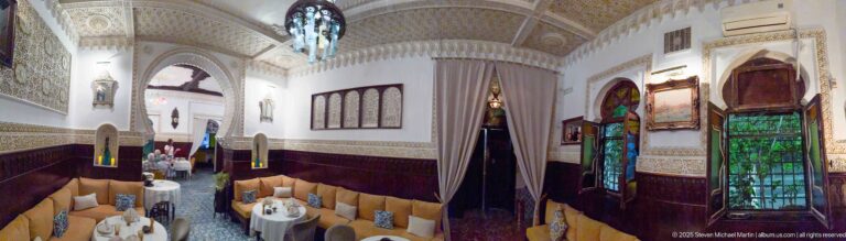 Ornate Algiers dining room with carved dark wood, patterned plasterwork, arched openings, colored glass, and upholstered seating.