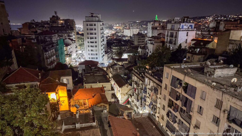 Night panorama of dense Algiers neighborhoods with apartment blocks, rooftops, illuminated streets, and scattered landmark lights.