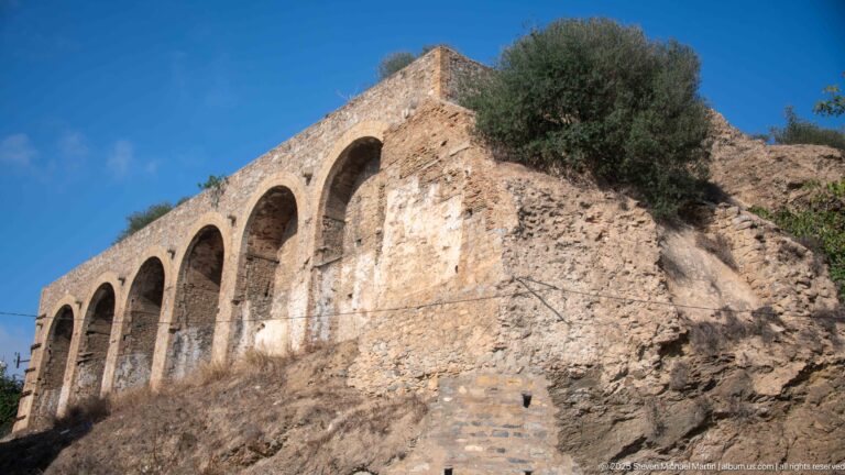 Historic stone-and-brick arcade with repeated tall arches built along a rocky hillside under blue sky.