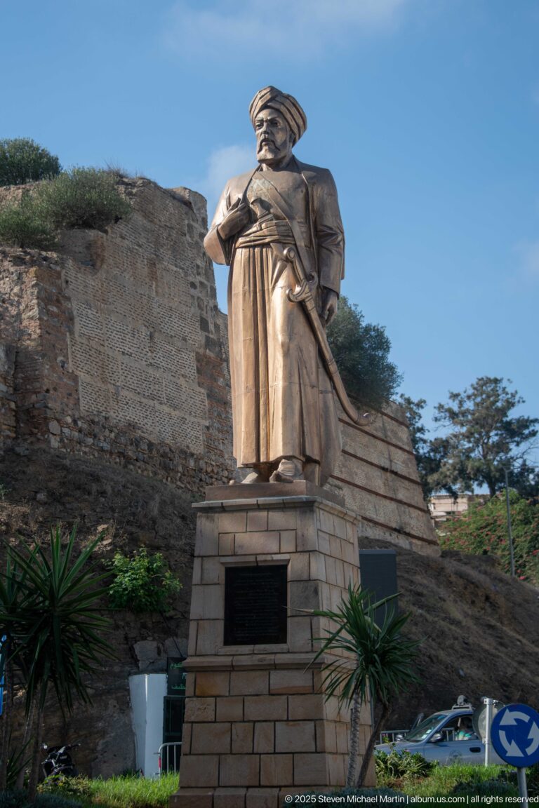 Bronze-colored statue of a robed man with turban-like headwear on a stone pedestal before old masonry walls.