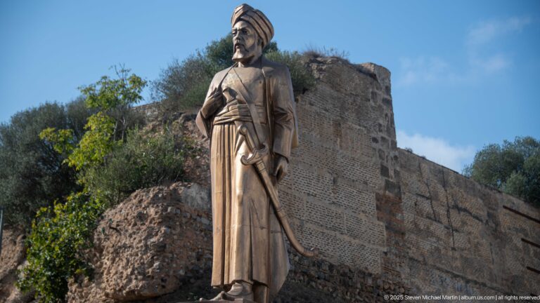 Side view of a robed commemorative statue with turban-like headwear before rough historic masonry and vegetation.