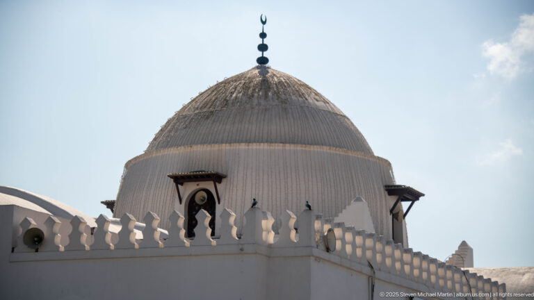 Close view of a ribbed white mosque dome with crescent finial, crenellated parapet, and small projecting windows.