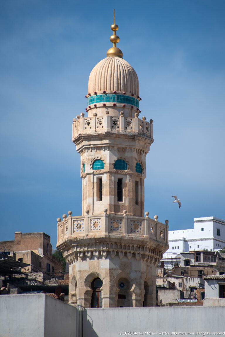 Ornate cylindrical minaret with balconies, turquoise accents, ribbed domed crown, and gold finial against blue sky.