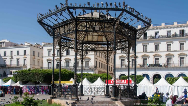 Ornate open-sided cast-iron pavilion with lattice roof in a busy Algiers square with tents and pedestrians.
