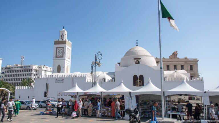 Algerian flag above a white mosque with square minaret and dome beside market tents and pedestrians.