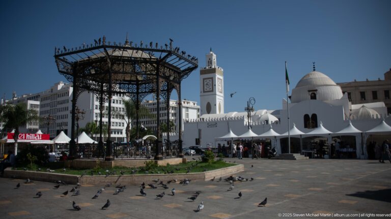 Public square in Algiers with ornate cast-iron pavilion, white mosque, clock-faced minaret, tents, pedestrians, and pigeons.