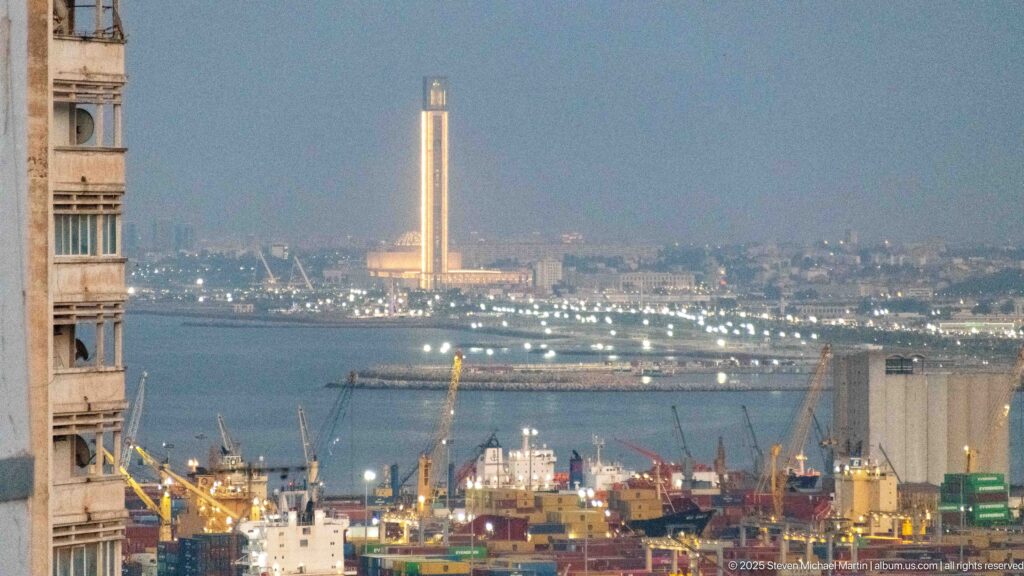 Algiers harbor at dusk with cranes, ships, waterfront lights, and a tall illuminated minaret across the bay.