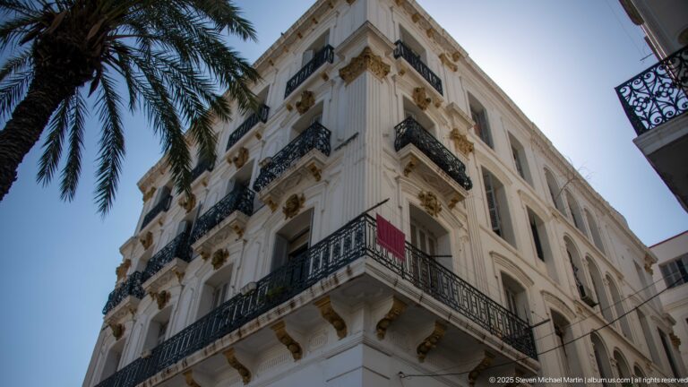 White corner building with black wrought-iron balconies, tall windows, and classical decorative details in Algiers.