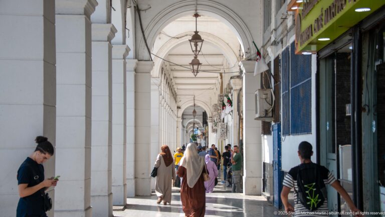 Pedestrians walking beneath a long white arcade with repeated arches, hanging lanterns, and storefronts in Algiers.