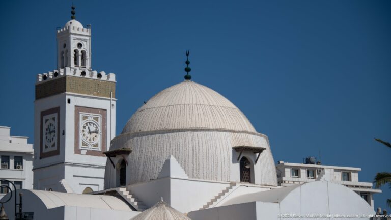 Close view of a ribbed white mosque dome beside a square minaret with clock faces and decorative panels.