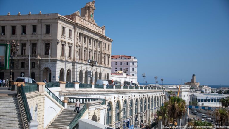 Large white civic building above broad stairs, arcaded terraces, palms, and the waterfront in Algiers.