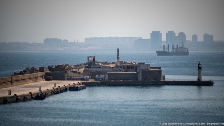 Algiers harbor entrance with breakwater lighthouse, low fortified buildings, cargo ship, and hazy skyline beyond.