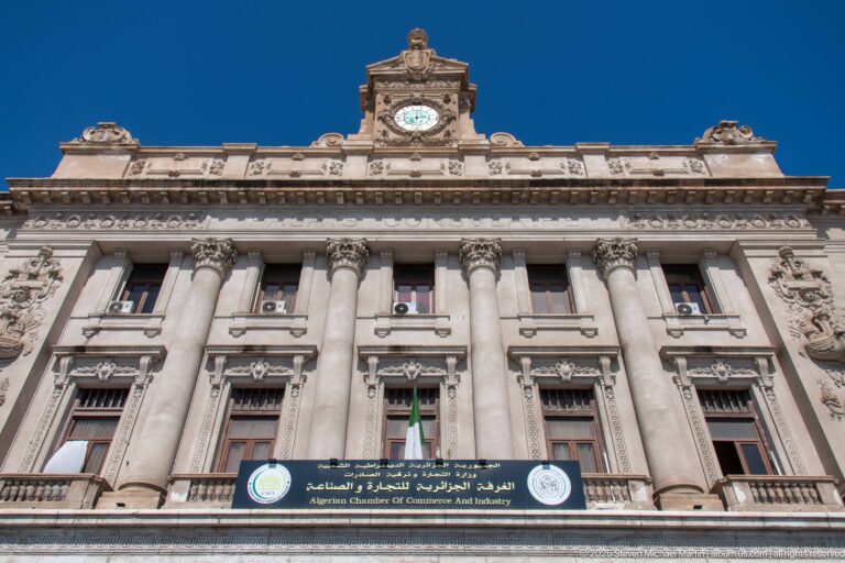 Grand stone façade of the Algerian Chamber of Commerce and Industry with columns, sculpted ornament, clock, and bilingual sign.