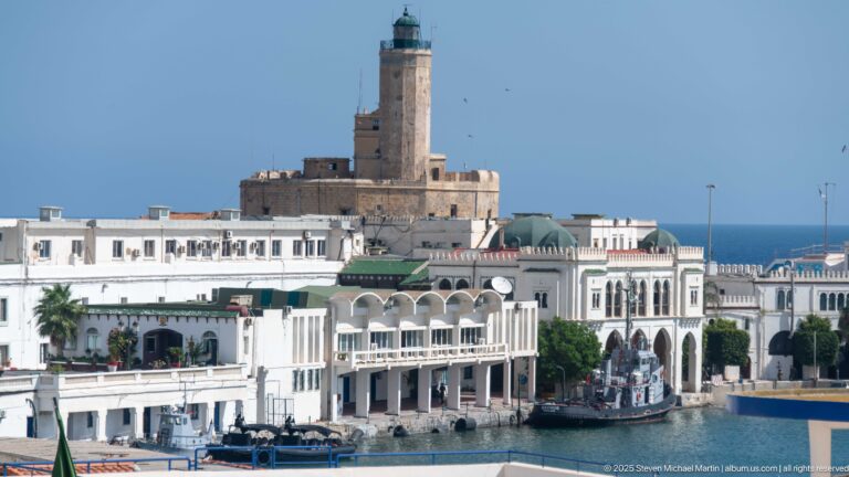Stone lighthouse tower above white arcaded waterfront buildings beside Algiers harbor.