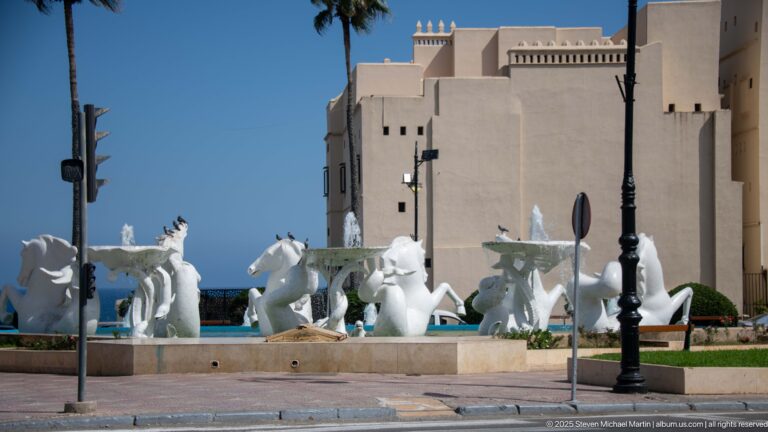 Group of stylized white horse sculptures in a fountain before a large pale civic building.