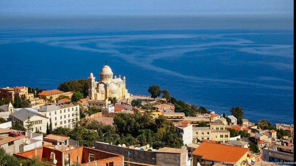 Notre-Dame d’Afrique on a hillside above red-roofed buildings and the deep blue Mediterranean Sea.