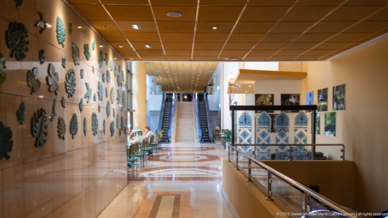 Polished hotel corridor with wall sculptures, blue-and-white patterned screens, and escalators at the far end.