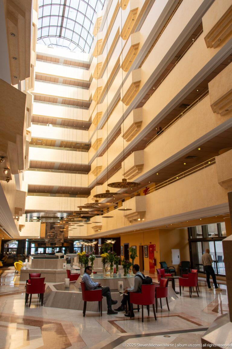 Wide hotel lobby with red chairs, polished floor, multiple balcony levels, plants, and bright atrium roof.