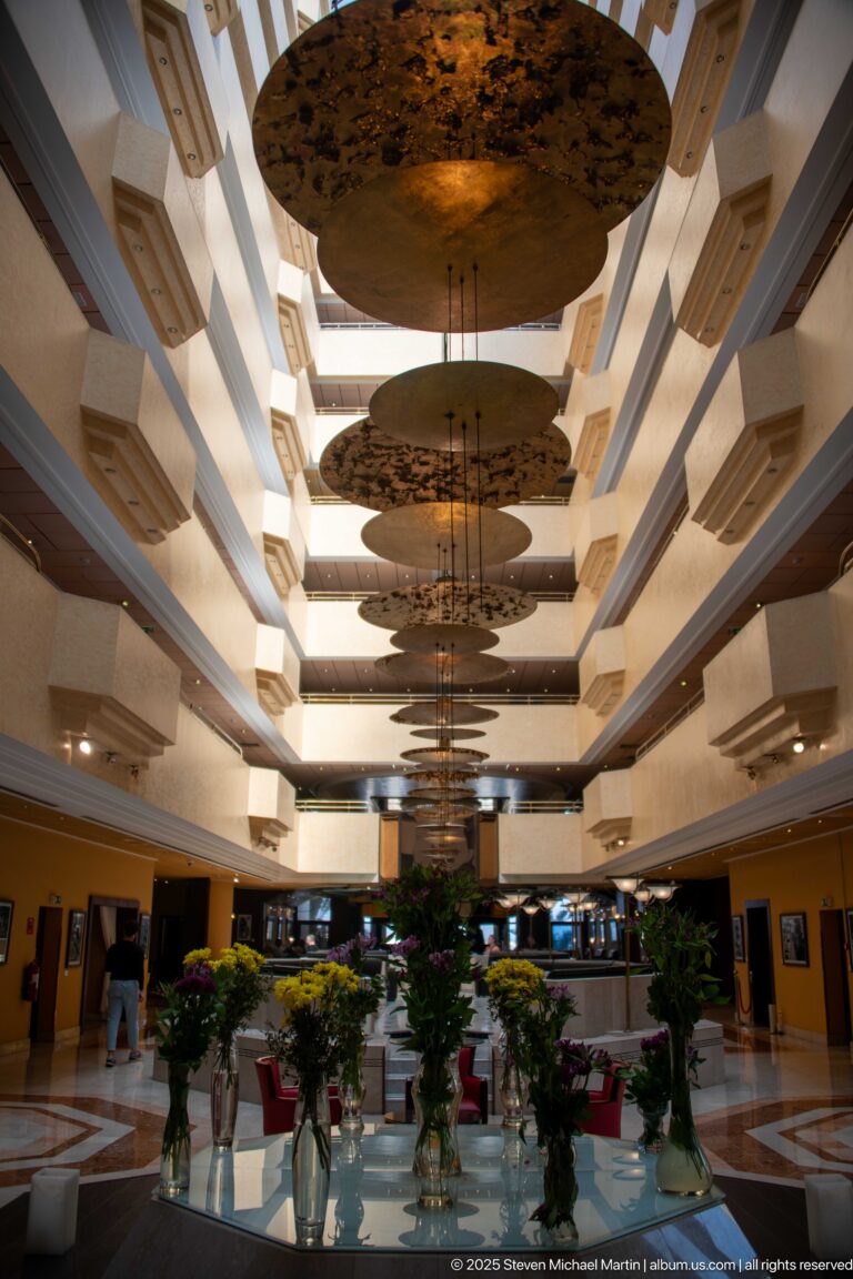 Multi-story hotel atrium with large suspended circular panels, central chandelier, flowers, and red lounge seating.