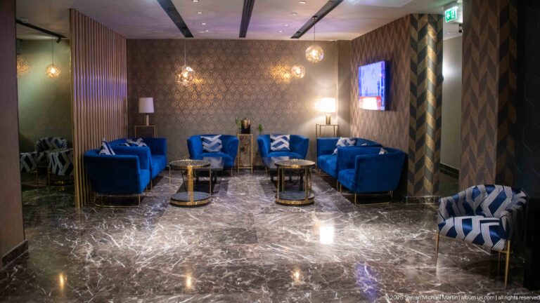 Hotel lounge with blue armchairs, round tables, patterned walls, pendant lights, and a polished dark stone floor.