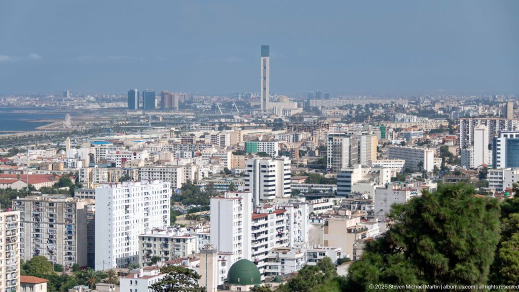 Elevated Algiers cityscape with dense neighborhoods and the tall Great Mosque minaret near the Mediterranean coast.