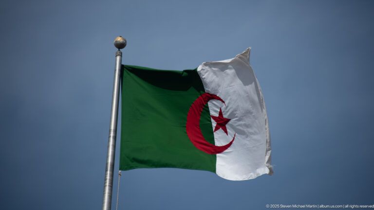 Algerian flag with green and white fields and red crescent and star flying on a tall pole.