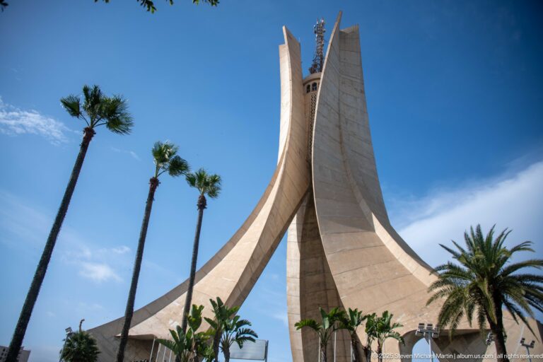 Martyrs’ Memorial rising above palm trees and landscaped grounds under blue sky.
