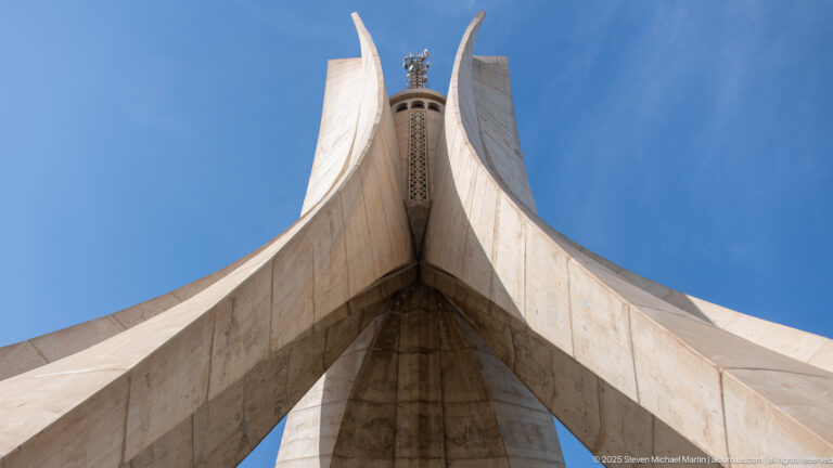 Low-angle view beneath the curving concrete fins of the Martyrs’ Memorial against a vivid blue sky.
