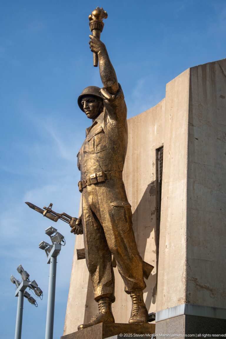Side view of a bronze-colored memorial figure with raised arm and weapon beside the Martyrs’ Memorial.