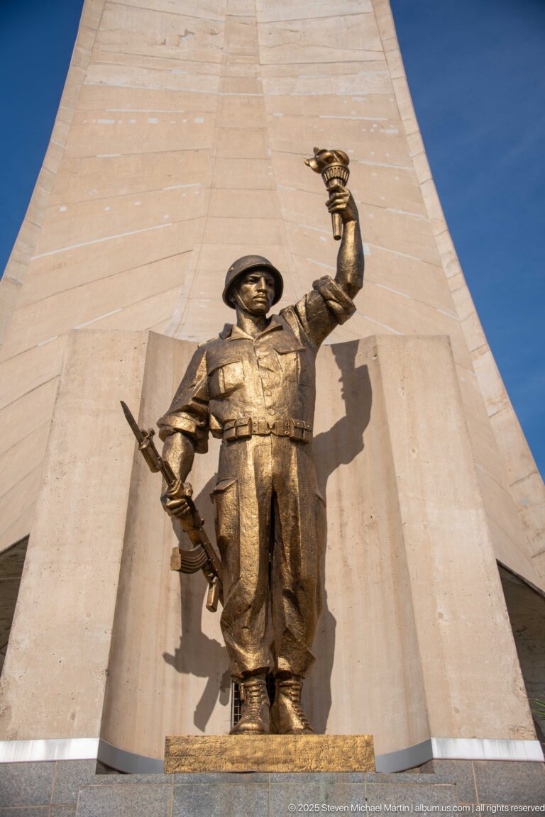 Front view of a bronze-colored memorial figure raising one arm high before the concrete Martyrs’ Memorial.