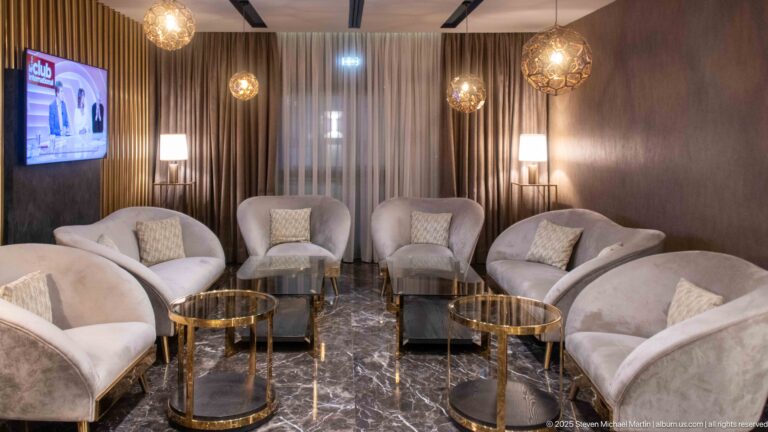 Contemporary hotel lounge with pale curved armchairs, brass-toned tables, curtains, lamps, and a polished dark floor.