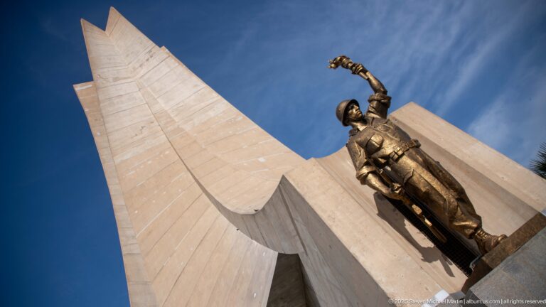 Bronze-colored memorial figure raising one arm beside a towering concrete fin of the Martyrs’ Memorial.