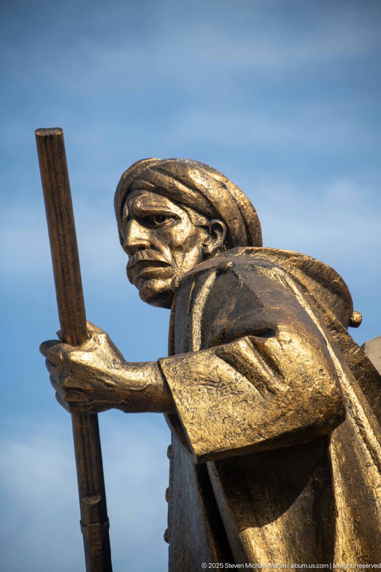 Close low-angle view of a bronze-colored memorial figure gripping a long vertical object against blue sky.