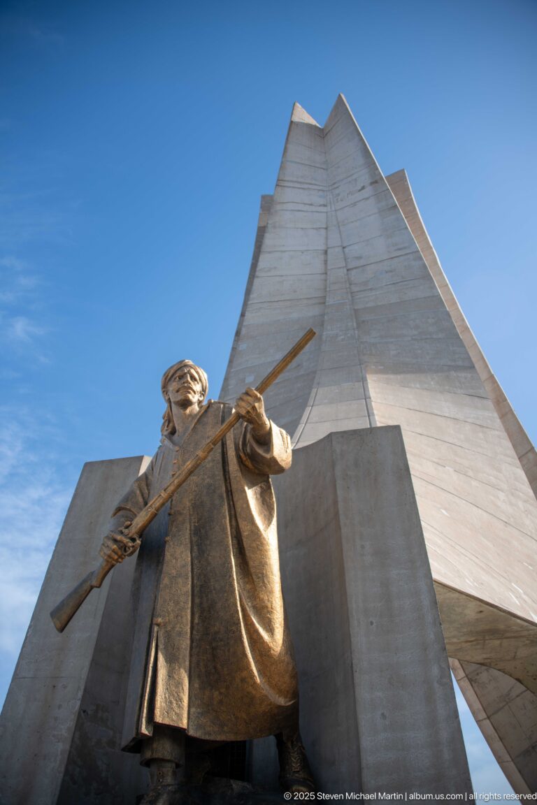 Bronze-colored standing figure holding a long weapon or staff before the concrete structure of the Martyrs’ Memorial.