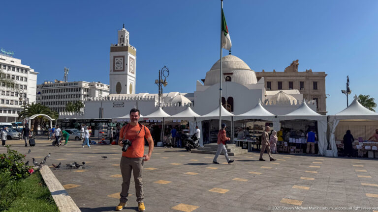 Man standing in an Algiers square with white market tents, mosque dome, Algerian flag, pedestrians, and pigeons.