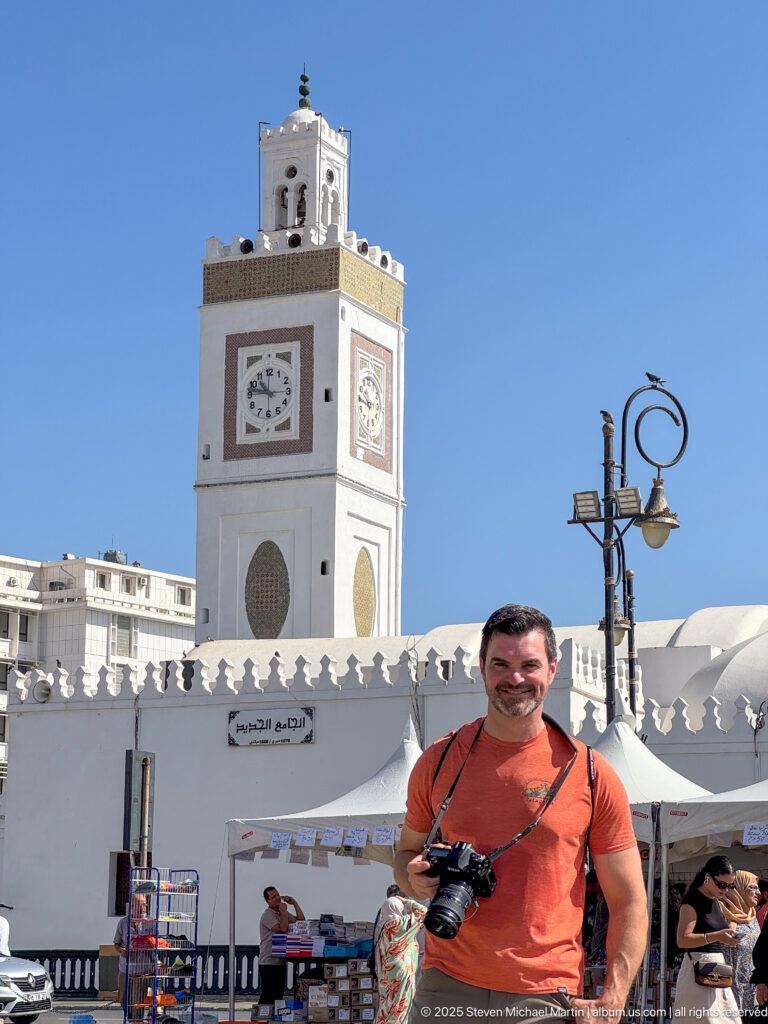 Man with camera standing before a white mosque and square clock-faced minaret in a busy Algiers square.