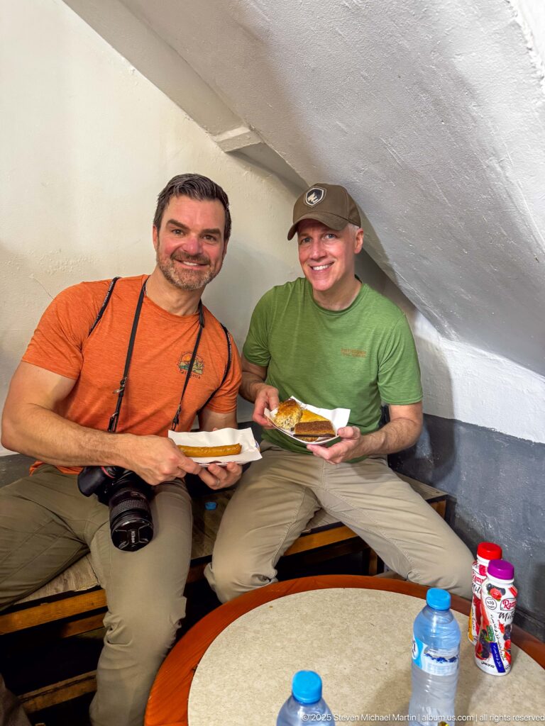 Two smiling men seated indoors holding small plates of food, with camera straps and water bottles nearby.