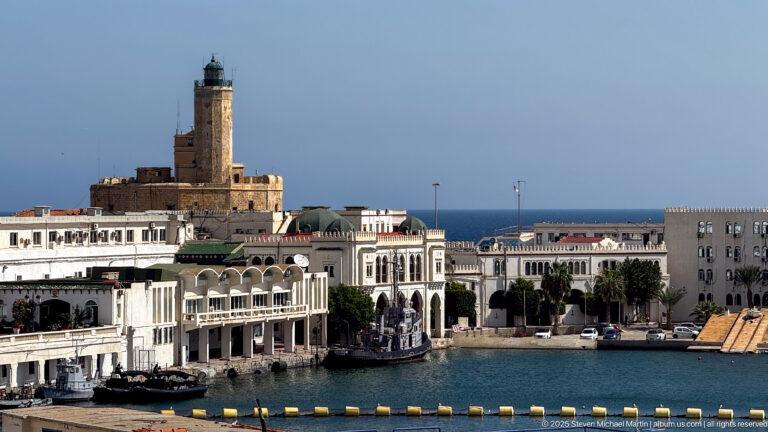 White harborfront buildings and arcades beneath a tall stone lighthouse tower beside blue water in Algiers.