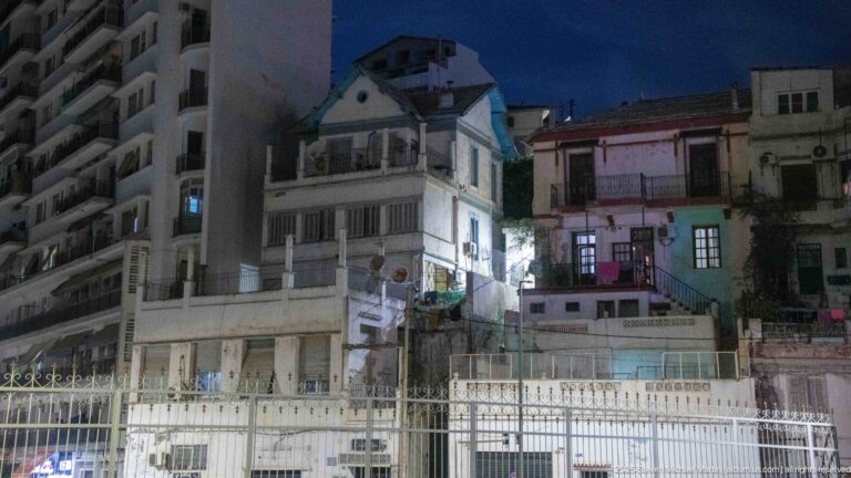 Night view of multi-story Algiers residential buildings with balconies and lit windows behind a long metal fence.