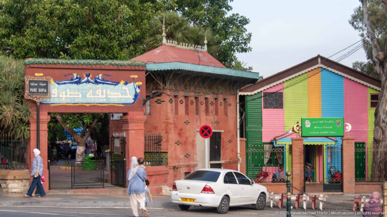 Arabic-signed park entrance beside a brightly painted multicolored wooden kiosk, with pedestrians and a parked car.