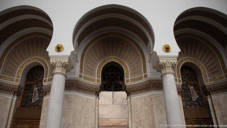 Close view of three recessed arches with pale columns, geometric decoration, calligraphic bands, doors, and dark grilles.