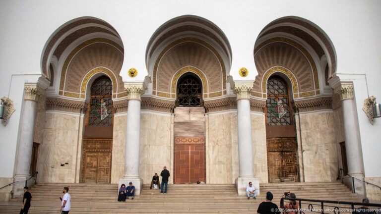 Three large arched portals with pale columns, carved ornament, wooden doors, and visitors on broad steps.