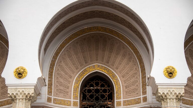 Large recessed arch with geometric ornament, gold Arabic calligraphy, dark grille, and decorative medallions.